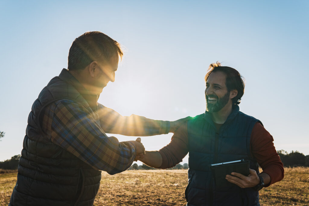Two happy farmers shaking hands in a cultivated field at sunset, making a business agreement