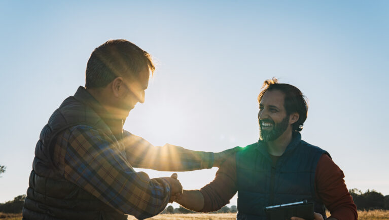 Two happy farmers shaking hands in a cultivated field at sunset, making a business agreement